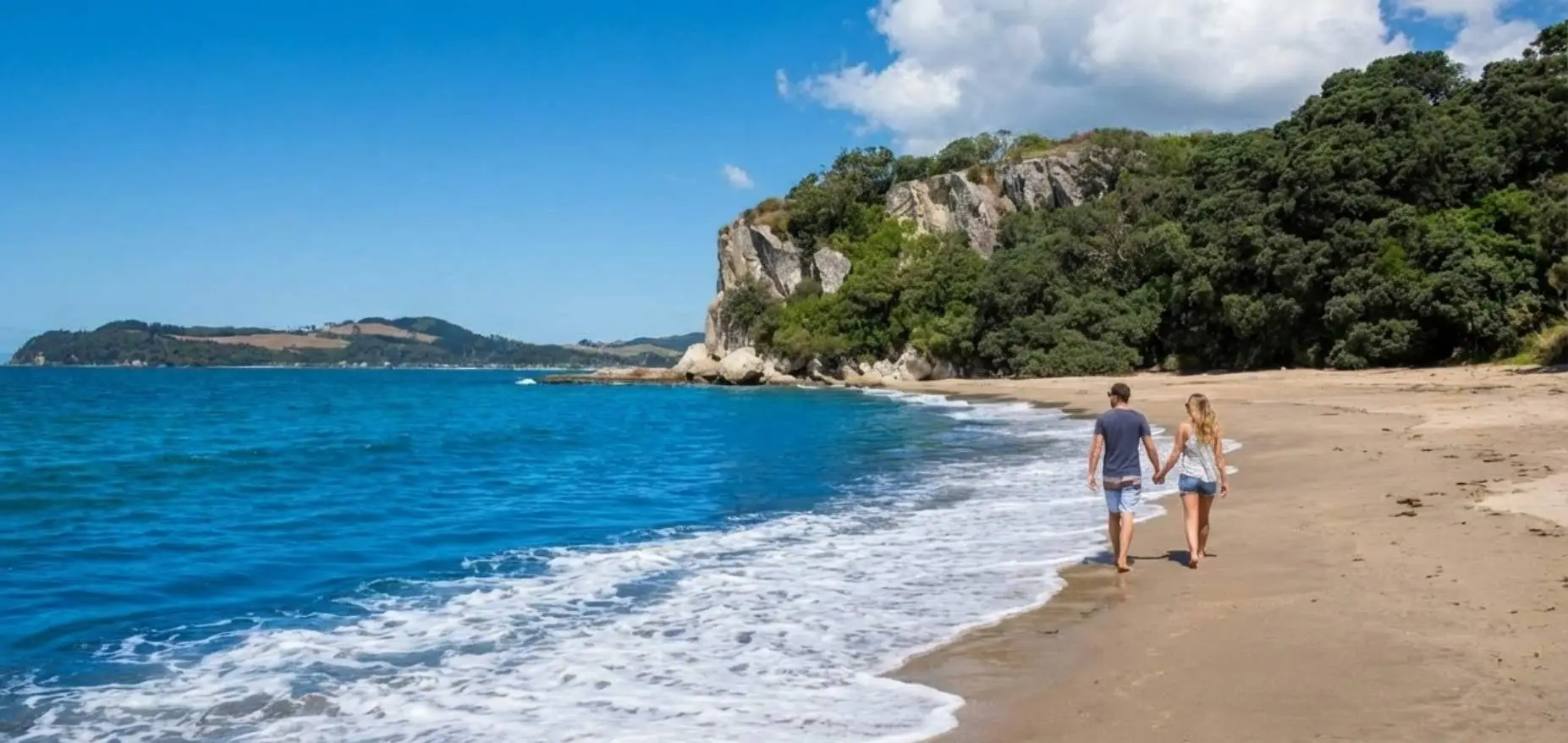Couple walking on Lonely Bay Beach with limestone cliffs and native bush behind them, Cooks Beach Coromandel