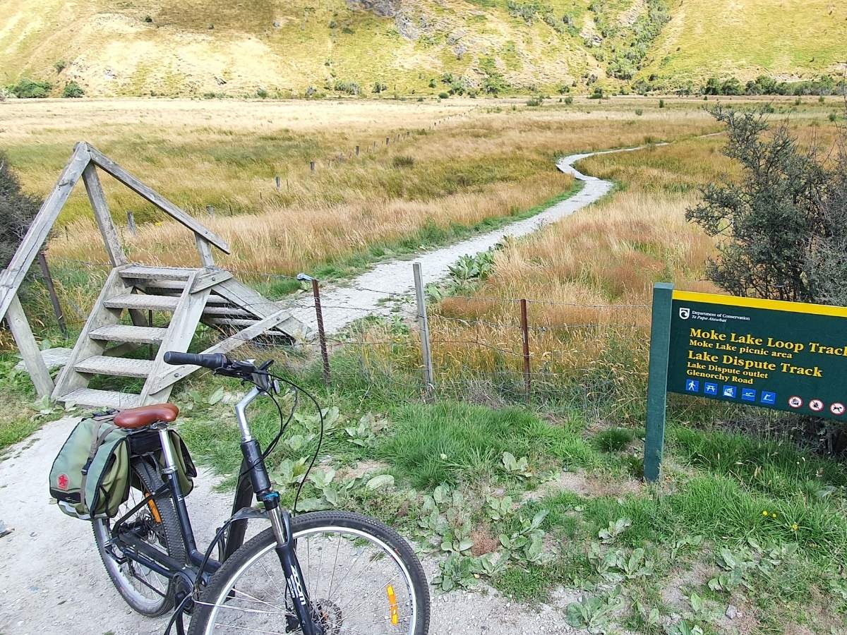 DOC sign and wooden stile at Moke Lake loop track with e-bike, showing track times for Moke Lake Loop and Lake Dispute tracks