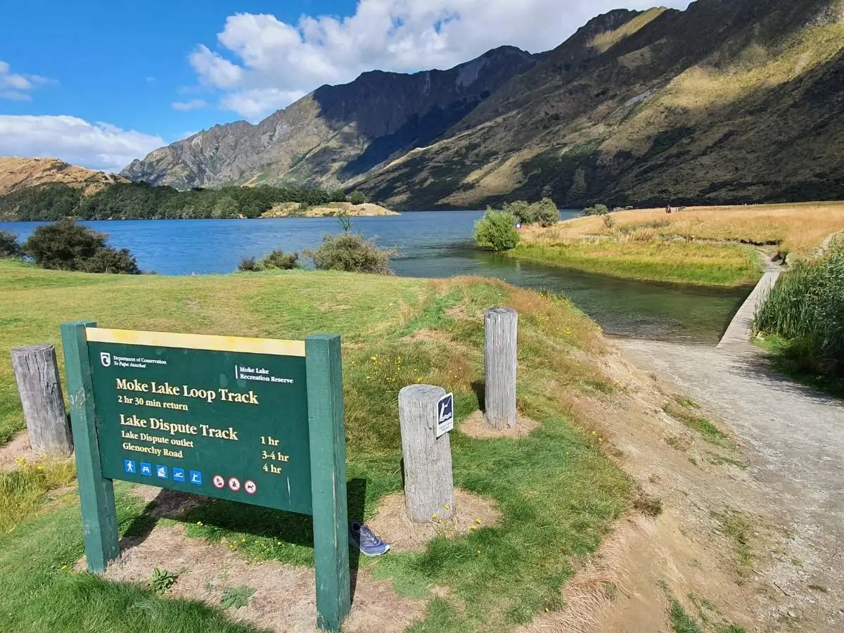 DOC sign at the start of Moke Lake Loop Track with lake and boardwalk visible behind, Queenstown