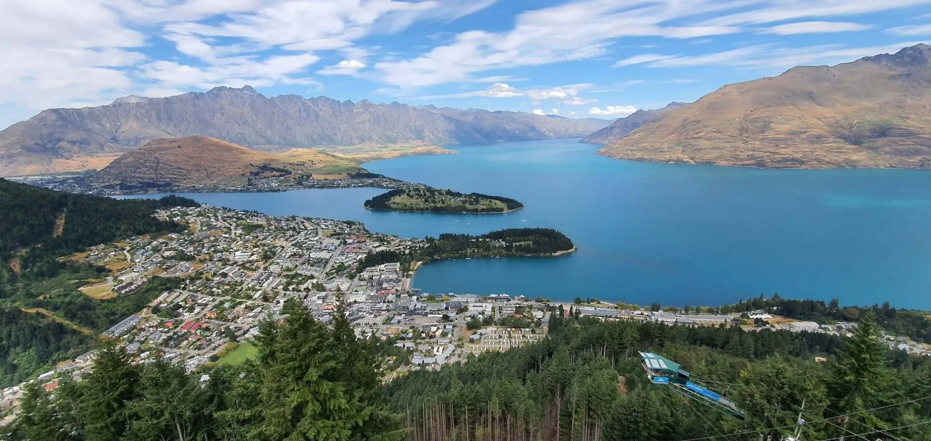 Panoramic aerial view of Queenstown town, Lake Wakatipu with peninsulas, Remarkables mountain range, and surrounding brown hills under blue sky