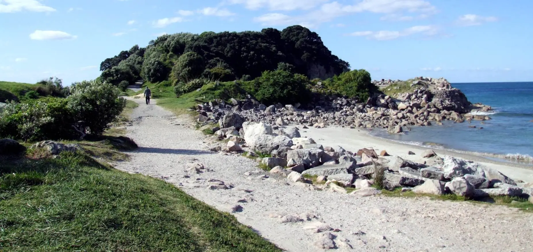 Walker on the gravel track approaching Moturiki Island (Leisure Island) at Mt Maunganui, with granite boulders and the Pacific Ocean alongside