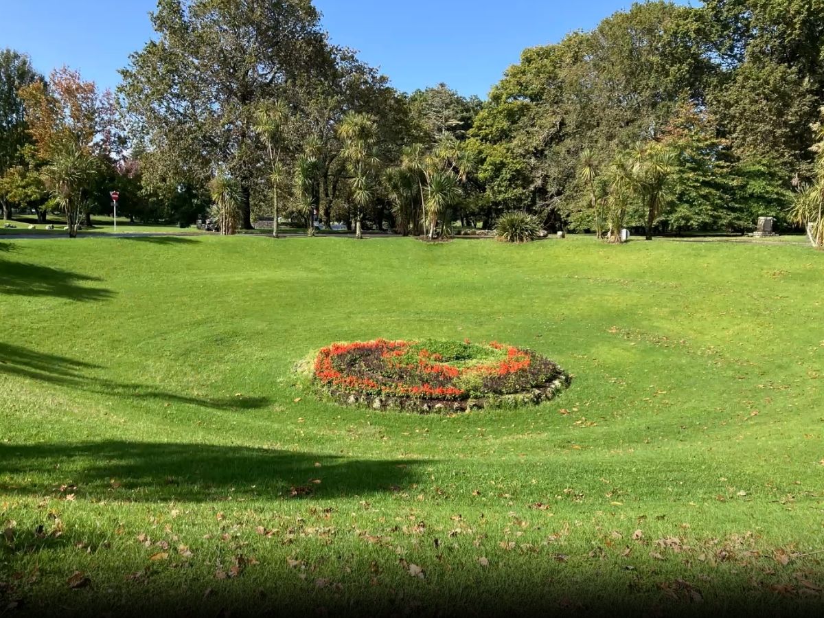 Circular sunken garden with red and green flower beds surrounded by green lawn, palm trees and mature trees in background