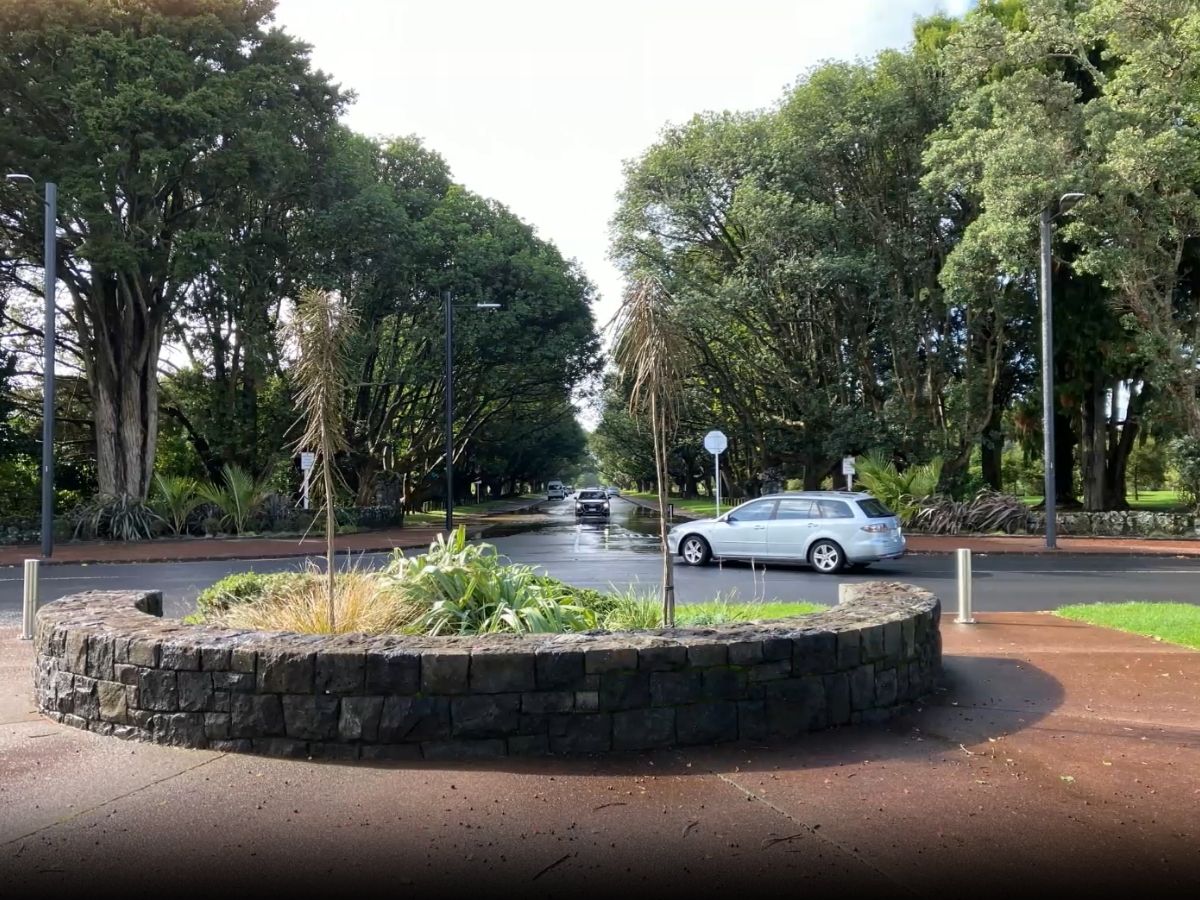 Wet sealed road lined with mature trees on both sides leading into Cornwall Park, stone planter in foreground