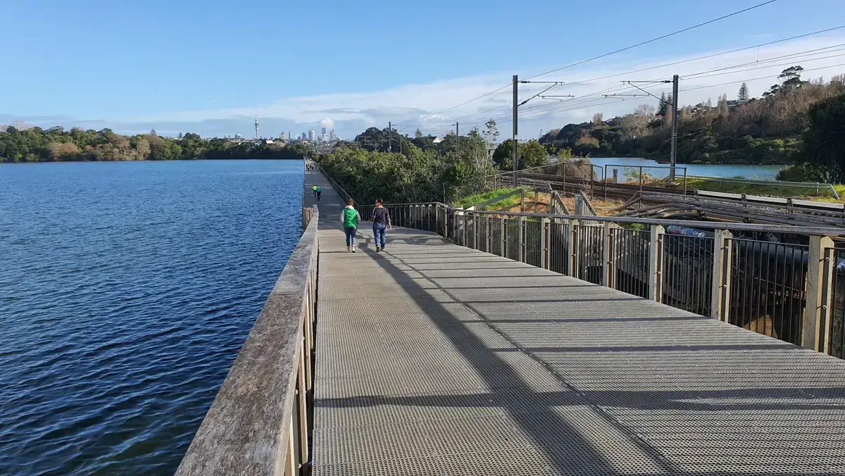Walkers on the Orakei Basin boardwalk beside the Eastern rail line with the Auckland city skyline in the distance